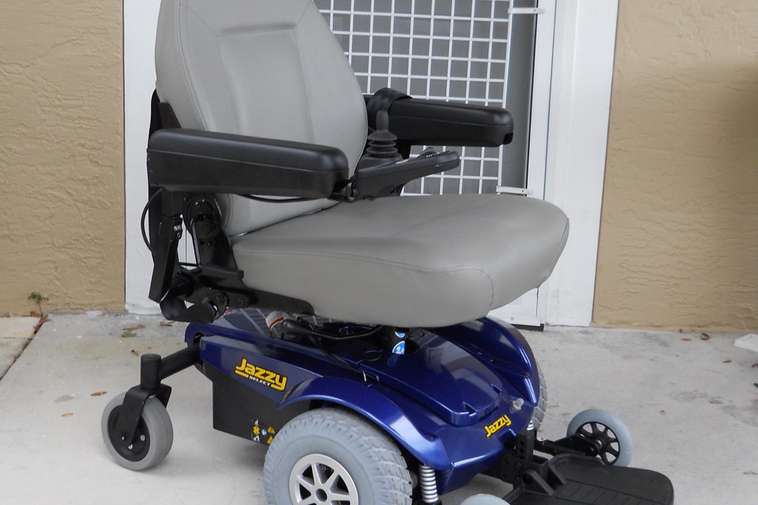 A blue Pride Jazzy Select power wheelchair with gray seat, joystick control, and mid-wheel drive configuration positioned on a home porch